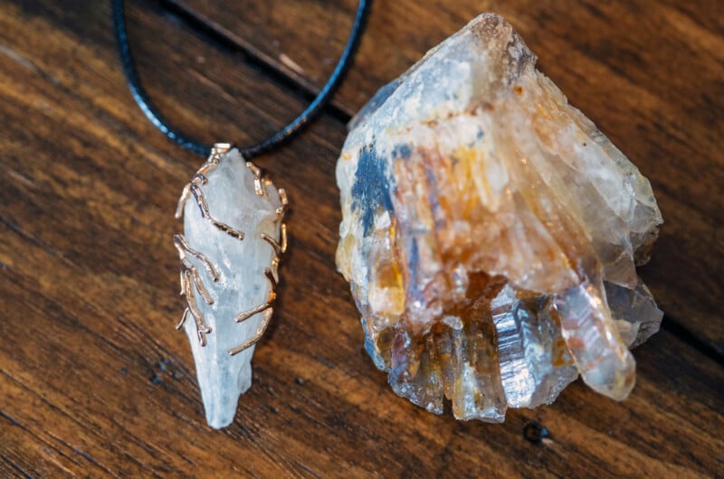 A clear quartz crystal pendant wrapped in wire lies next to a rough, multicolored quartz mineral on a wooden surface.