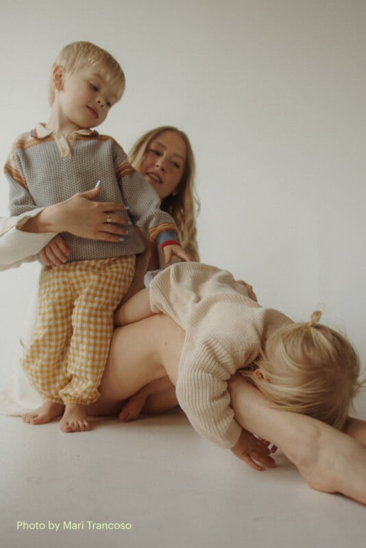 A woman sits on the floor, holding two young children. One child in yellow checkered pants stands near her, while the other, dressed in beige, leans across her legs in a playful pose. The background is neutral and minimal.