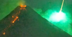 A volcano erupts at night, with streams of glowing lava flowing down its side and smoke rising, while a bright bolt or flash of light appears in the green-tinged sky nearby.