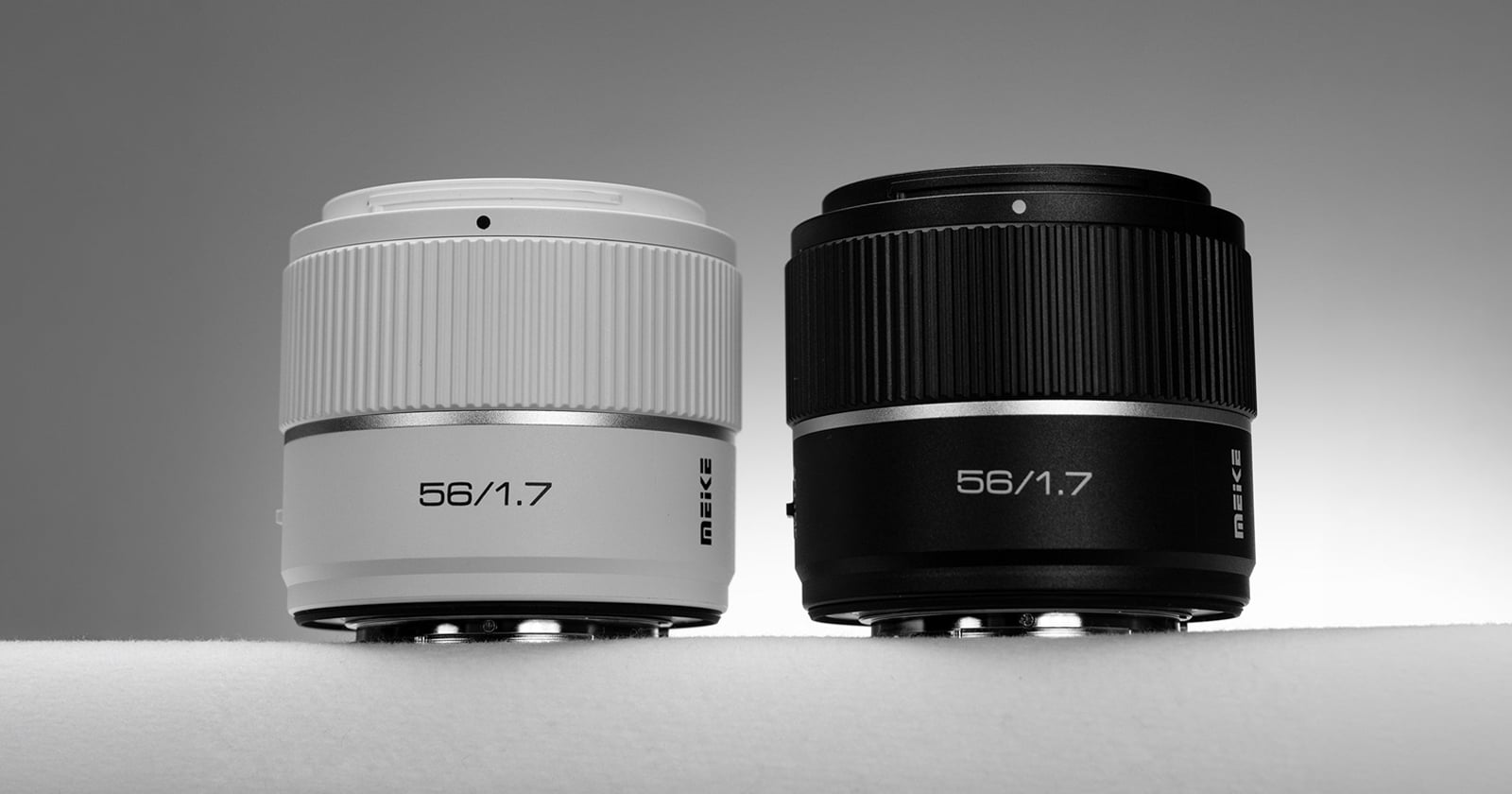 Two camera lenses are displayed side by side against a neutral background, one in white and the other in black. Both lenses have "56/1.7" prominently printed on the front.