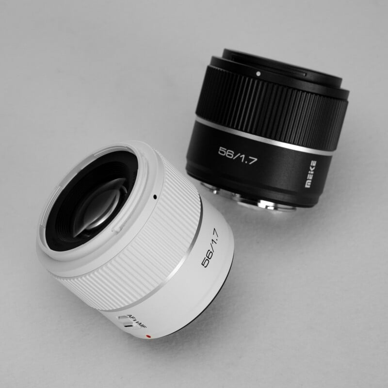 Two camera lenses with “56/1.7” markings, one white and one black, are displayed on a light grey background. Both lenses are from the brand Meike and are shown at an angle.