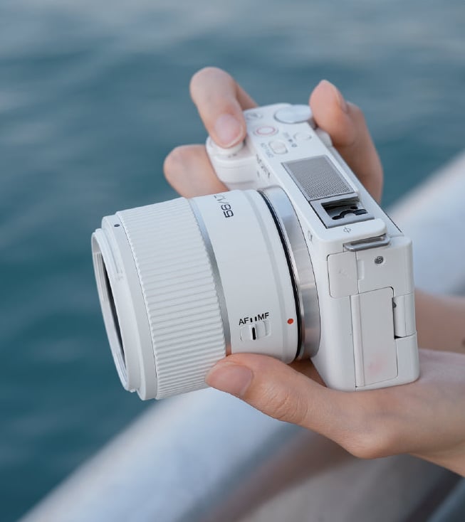 A person holds a white digital camera with both hands near a body of water, focusing on adjusting the lens.