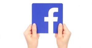 Two hands hold a blue square card featuring the white lowercase "f" Facebook logo against a plain white background.