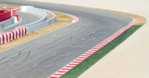 A curved section of a racetrack with red and white striped curbs, tire marks on the asphalt, and protective barriers along the side. The surrounding areas are sandy and grassy.