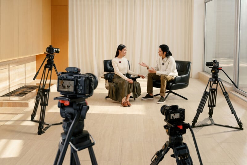 Two people sit and talk in armchairs in a bright room, surrounded by three video cameras on tripods, suggesting an interview or filmed conversation.