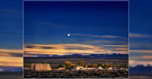 Adobe church and graveyard under a twilight sky with a full moon and snow-capped mountains in the distance.