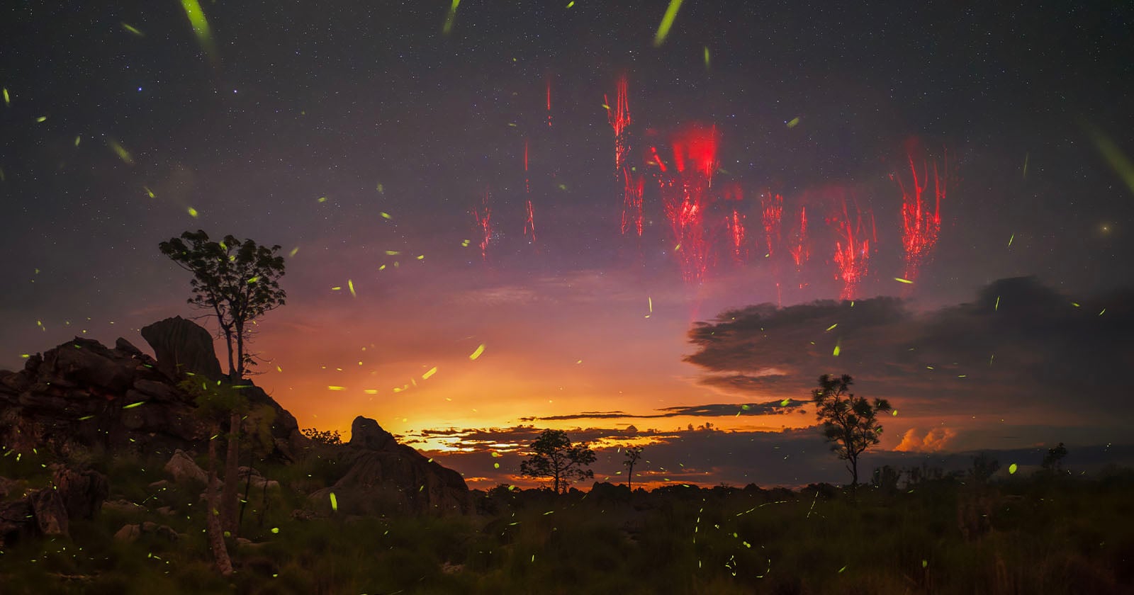 A dramatic sunset sky over a rocky landscape with scattered trees, glowing fireflies, and streaks of red sprites illuminating the night clouds and starry sky above.