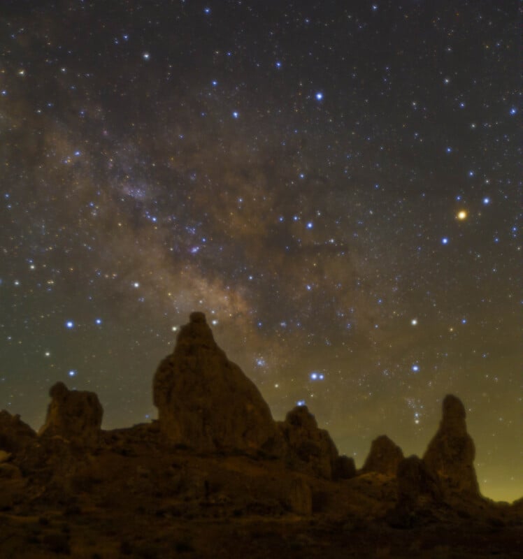 Jagged rock formations are silhouetted against a night sky filled with countless stars and the bright, cloudy band of the Milky Way galaxy stretching across the background.