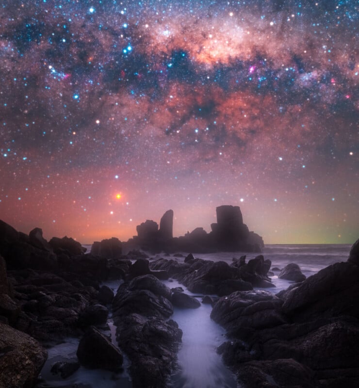 Rocky coastline at night with dark jagged rocks in the foreground and a vibrant, colorful Milky Way galaxy stretching across the star-filled sky above the horizon.