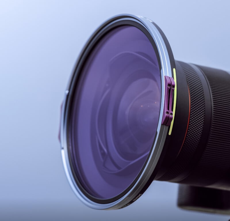 Close-up of a camera lens with a large, round, purple-tinted filter attached to the front, against a plain light background. The filter is secured with small purple clips.