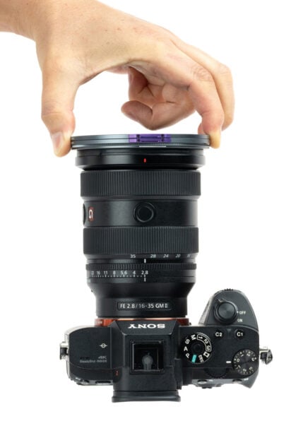 A hand is attaching a round camera lens filter to the front of a Sony camera with a mounted FE 2.8/16-35 GM lens, viewed from above on a white background.