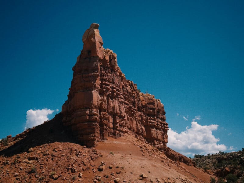 A tall, rugged red rock formation rises from a dry, reddish-brown hillside under a clear blue sky with a few white clouds in the background. Sparse vegetation is visible at the base of the formation.