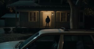 A person stands alone on the porch of a dimly lit house at night, partially silhouetted by the glow from the doorway, with a white car parked in the foreground.