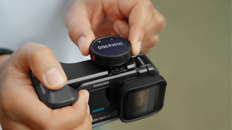 A person attaches a small round Insta360 accessory to the top of an Insta360 camera, holding the device with both hands. The camera is black with a large lens and a touchscreen.