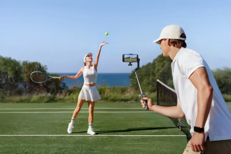 A woman in a white tennis outfit serves a tennis ball on an outdoor court, while a man in white records her with a smartphone on a gimbal. Trees and the ocean are visible in the background.