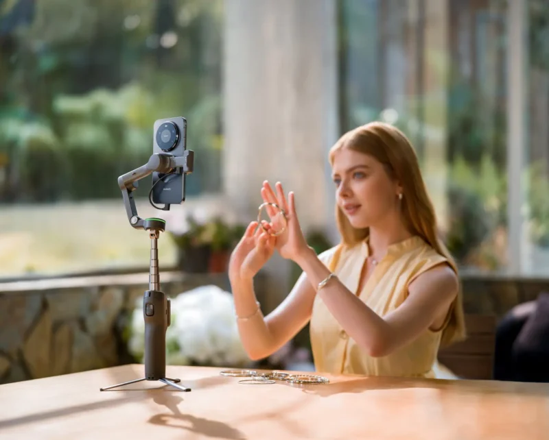 A woman sits at a table using a smartphone mounted on a tripod to record herself as she displays jewelry on her hands. Sunlight streams in through large windows, creating a bright, airy atmosphere.