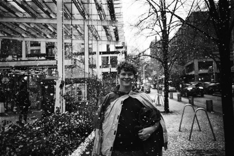 A young person stands smiling on a city sidewalk in snowfall, wearing a jacket draped over their shoulders. Leafless trees, parked cars, and buildings with large windows line the street. The scene is in black and white.