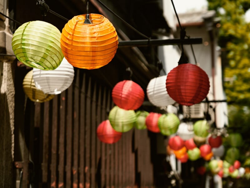 Colorful paper lanterns in green, orange, red, and white hang in a row outside a building, creating a festive and vibrant atmosphere. The background is slightly blurred, emphasizing the lanterns' details.