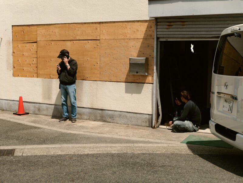 A man in a black jacket and cap stands outside using his phone, while another man sits on the ground inside a partially open garage, gesturing with his hand. A white van and an orange traffic cone are nearby.