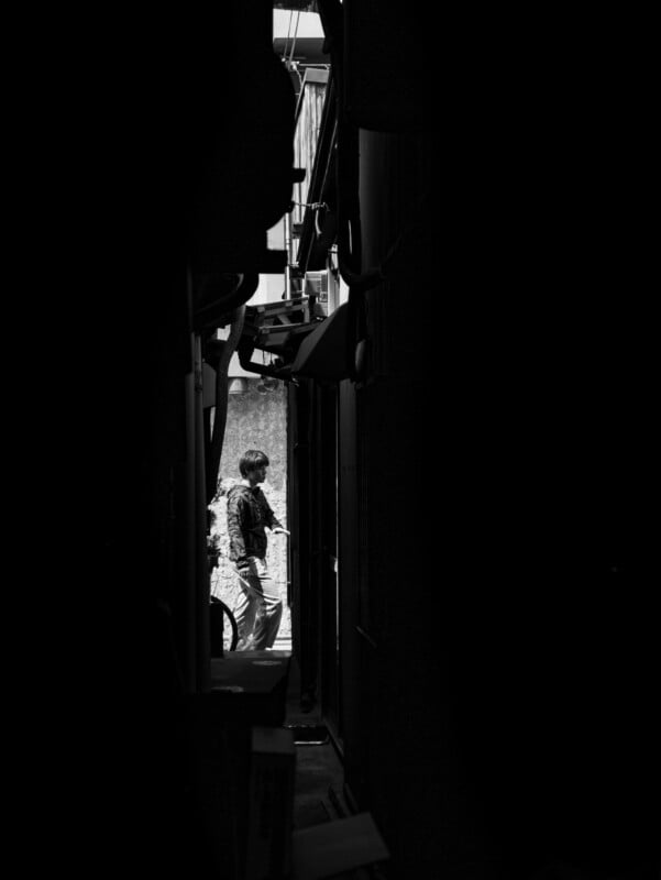 A person walks through a narrow alleyway, illuminated by daylight, while the surrounding area is in deep shadow. The scene is in black and white, highlighting strong contrasts and textures.