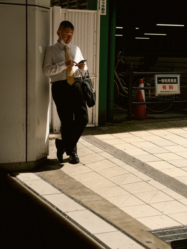 A man in business attire leans against a wall in sunlight, looking at his phone and holding a black bag. Bicycles, traffic cones, and a sign are visible in the shaded background.