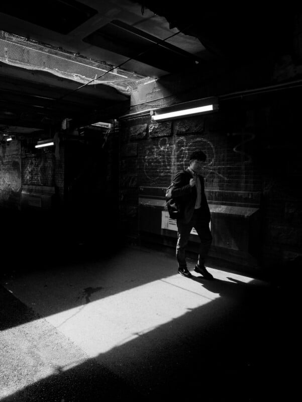 A man stands alone in a dimly lit urban underpass, with sunlight casting a sharp shadow on the ground. He carries a bag over his shoulder and looks down, surrounded by graffiti-covered walls.