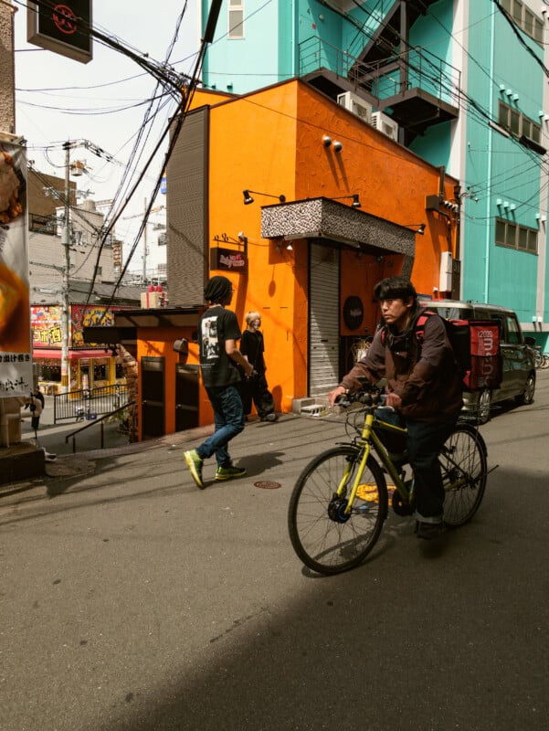 A person rides a bicycle past an orange building on a city street while two others walk nearby; colorful urban scene with overhead wires and various signs visible.