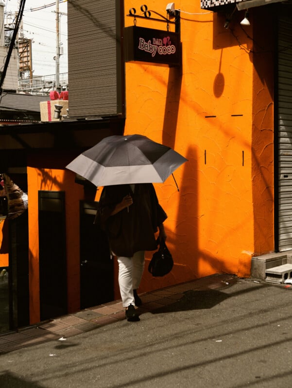 A person holding a large black-and-white umbrella walks up a sloped street past a bright orange building with a sign that reads "Bar Baby coco." Shadows and power lines are visible on the wall.