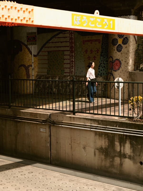A woman walks along a sunlit train platform with colorful mosaic art on the wall behind her and a yellow sign overhead, partially written in Japanese. A black fence separates the platform from the tracks below.
