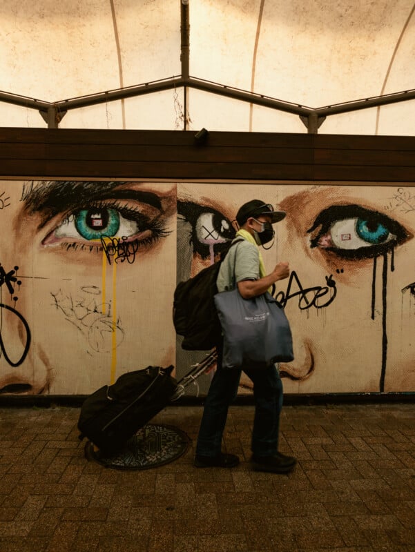 A person wearing a mask and cap walks past a mural of large, realistic blue eyes on a wall. They carry a tote bag and pull a suitcase, while graffiti surrounds the mural in a covered walkway.
