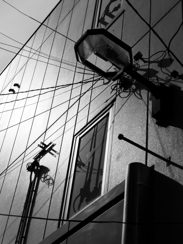 A black-and-white photo of a building exterior with a streetlight, visible power lines, cables, and their shadows intersecting across a reflective wall and window.