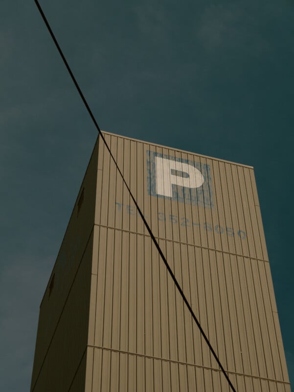 A tall beige building with a large blue and white “P” sign, indicating parking, stands against a cloudy sky. A black cable runs diagonally across the image.