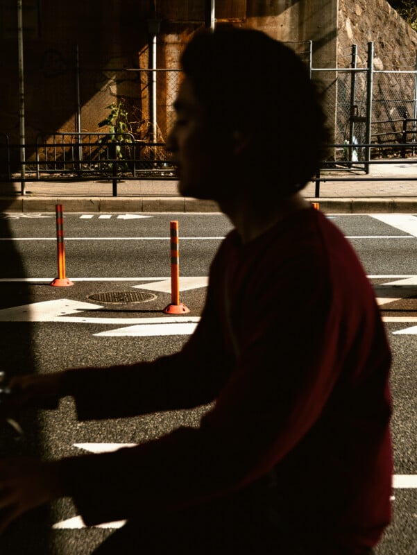 A person in a dark red shirt is captured in silhouette, riding a bicycle on a street with orange traffic posts and road markings, with sunlight and shadows creating strong contrast.