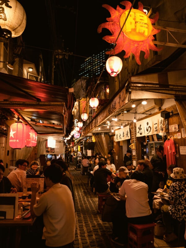 A lively, narrow street at night with people dining at outdoor tables under glowing red and white lanterns, surrounded by signs and banners in Japanese. The scene is warm and bustling with vibrant energy.
