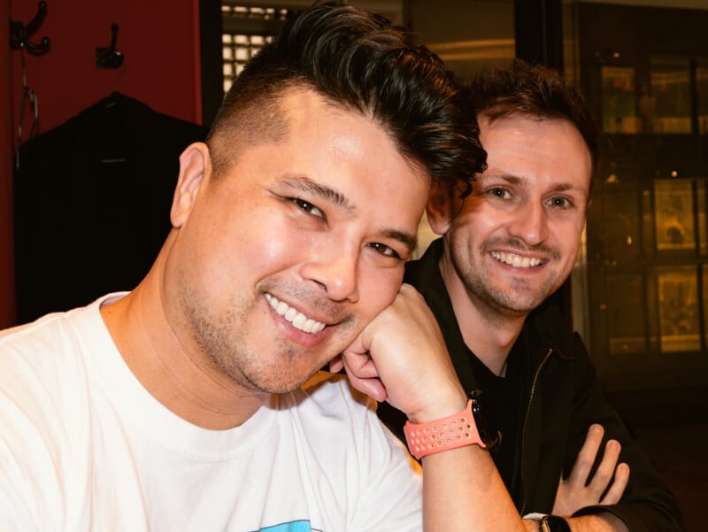 Two smiling men sit close together indoors. The man in front wears a white T-shirt and a smartwatch with a pink strap, resting his chin on his hand. The man behind wears a black shirt. Both look toward the camera.