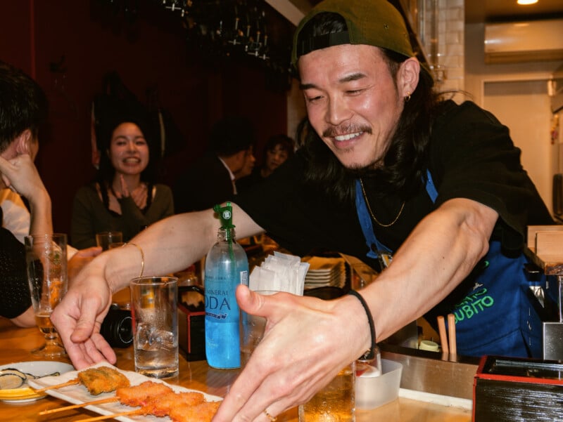 A smiling man in a cap and apron serves skewered fried food to a customer at a busy restaurant counter, with drinks and plates on the wooden bar and other diners in the background.