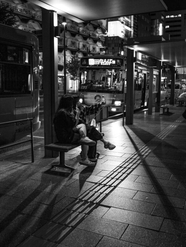 Two people sit on a bench at a bus stop at night, illuminated by streetlights. A bus is stopped in the background, and city buildings with lit windows are visible. The scene is in black and white.