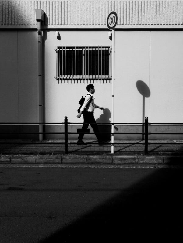 A person with a backpack walks past a white building casting strong shadows, with a barred window and a 30 km/h speed limit sign above. The scene is in black and white.