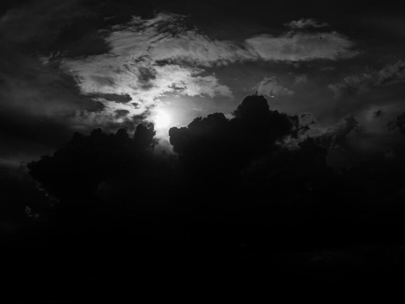 A dramatic black and white photo of the sun partially hidden behind large, dark clouds, with light rays streaming through openings in the cloud cover.