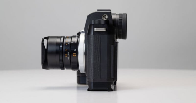 A black digital camera with a lens attached is shown in profile view, positioned on a white surface against a light gray background.