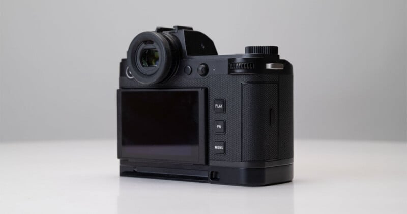 A black digital camera is positioned on a white surface, shown from the back with a viewfinder, a screen, and several buttons labeled "PLAY," "FN," and "MENU." The background is plain and light gray.