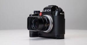 A black Leica camera with a lens attached is placed on a white surface, shown against a light gray background. The camera is angled slightly to the left, displaying the LEICA logo prominently.