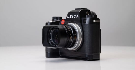A black Leica camera with a lens attached is placed on a white surface, shown against a light gray background. The camera is angled slightly to the left, displaying the LEICA logo prominently.