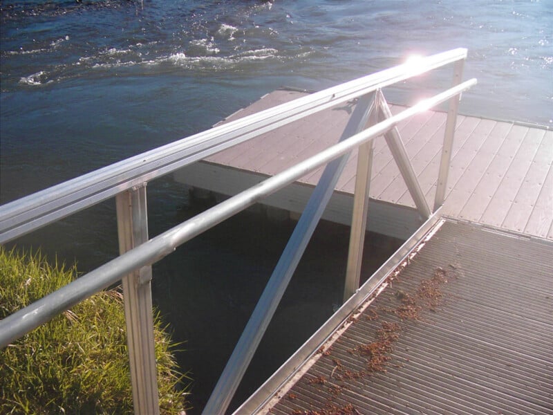 A metal ramp with a railing leads down to a floating dock on a body of water, with sunlight reflecting off the metal and dock surface. Some grass and scattered debris are visible on the ramp.