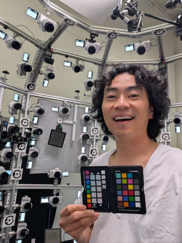 A person with curly dark hair smiles while holding a color calibration card in front of a large array of cameras and equipment, likely in a photo or 3D scanning studio.
