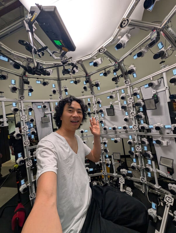 A person in a white shirt takes a selfie while sitting inside a circular structure lined with numerous cameras and lights, likely used for 3D imaging or motion capture. The person is smiling and waving.