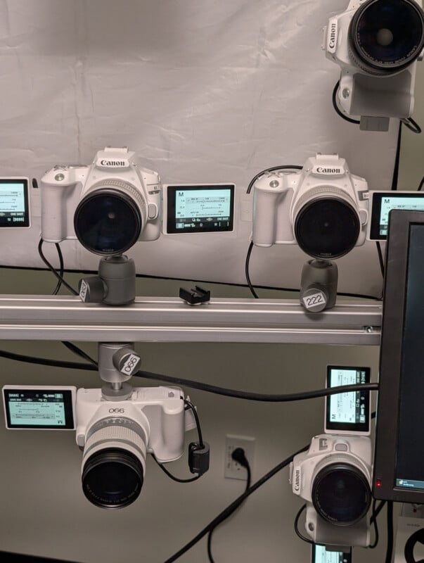 Several white Canon cameras are mounted on a metal rail, each with its LCD screen displaying camera settings. A monitor is partially visible on the right, and a power outlet is seen on the wall in the background.