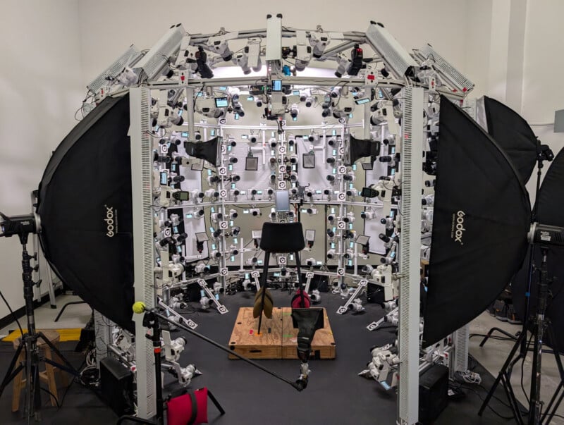 A chair is placed at the center of a large circular rig filled with multiple cameras and lights, surrounded by equipment in a studio setting. Two large Godox softboxes flank the setup.