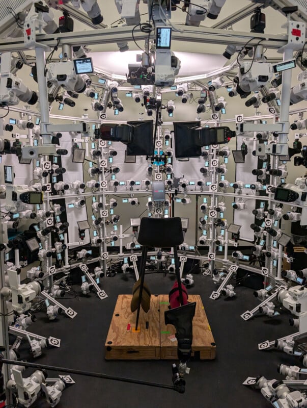 A circular rig with dozens of mounted cameras and lights surrounds a chair on a wooden platform, likely used for 3D imaging or photogrammetry in a studio setting.