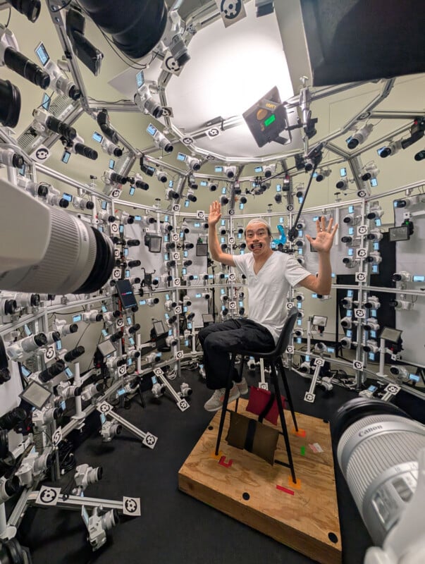 A man sits on a chair in the center of a room surrounded by numerous cameras and lights arranged on a circular frame, raising his hands and smiling at the camera.